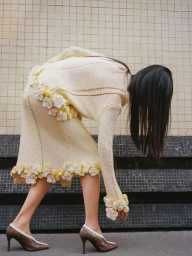 Model bending forward wearing a cream knit dress with floral appliqués and brown heels against a tiled wall