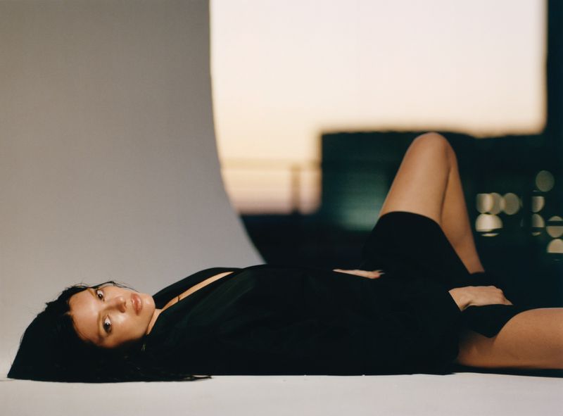 A young woman with dark hair lies on her side wearing a dark jacket with blurred city lights in the background, in a relaxed pose.