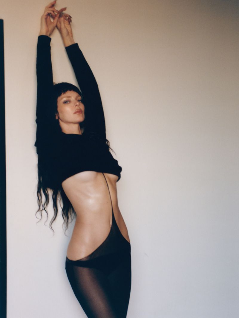 Adult female in black crop top and sheer maxi dress with arms raised, posing indoors against neutral wall in a fashion shot.