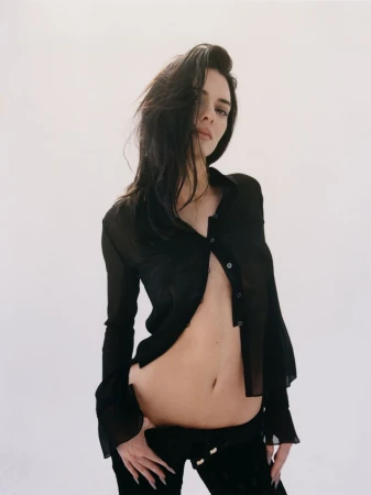 Brunette woman in sheer button-down shirt and dark pants posing against a neutral backdrop, showcasing a fashion-forward aesthetic.