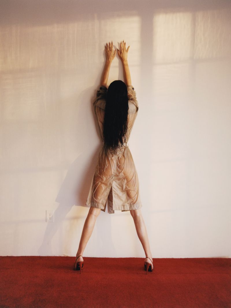 Back view of adult woman with long black hair, tan dress, and red heels, pressed against a white wall with hands raised on a red carpet.