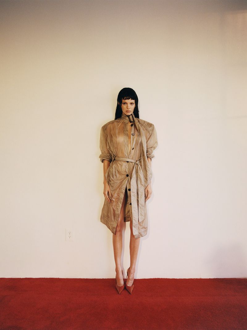 A long-haired woman in a sheer tan trench coat, red heels poses in front of a cream-colored wall with a red floor in a studio shot.