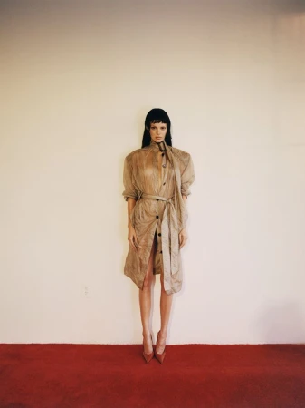 A long-haired woman in a sheer tan trench coat, red heels poses in front of a cream-colored wall with a red floor in a studio shot.