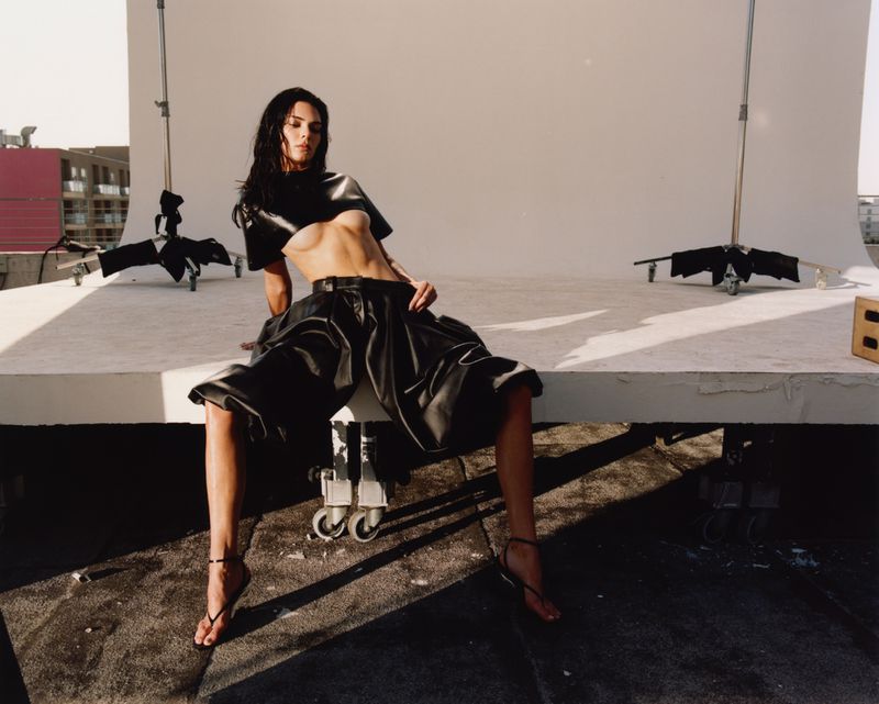 Adult woman with wet hair poses in black top and wide shorts with black sandals on a white stage with studio lighting equipment.