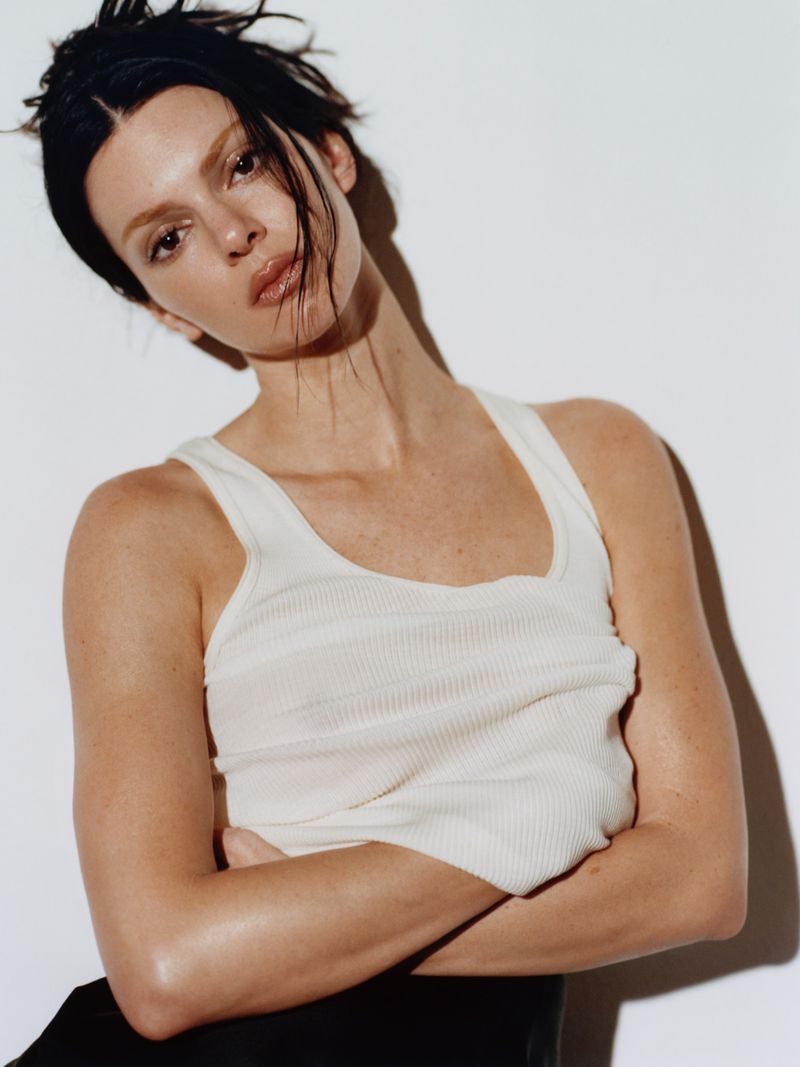 Indoor portrait of a woman with dark hair wearing a light tank top, with her arms crossed in front of a white background.