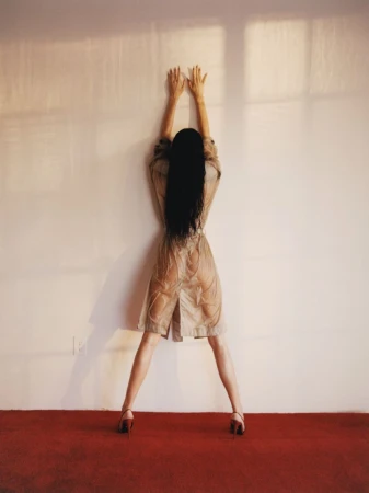 Kendall Jenner facing a plain white wall with arms raised, wearing a beige trench coat and heels on a red carpet