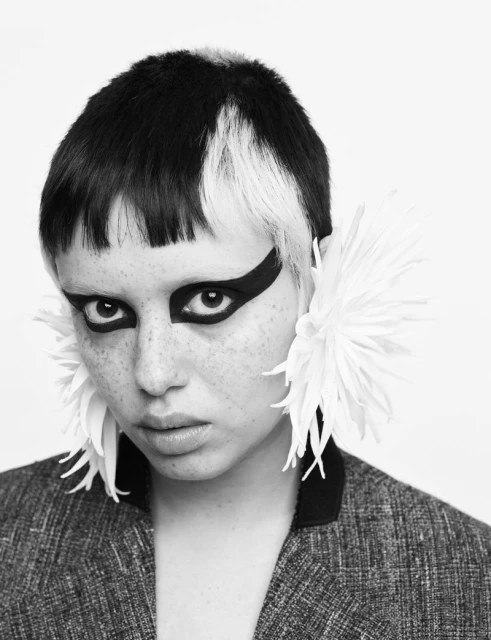 Monochrome portrait of a young adult female with white and black hair, exaggerated black eye makeup, and large flower earrings.