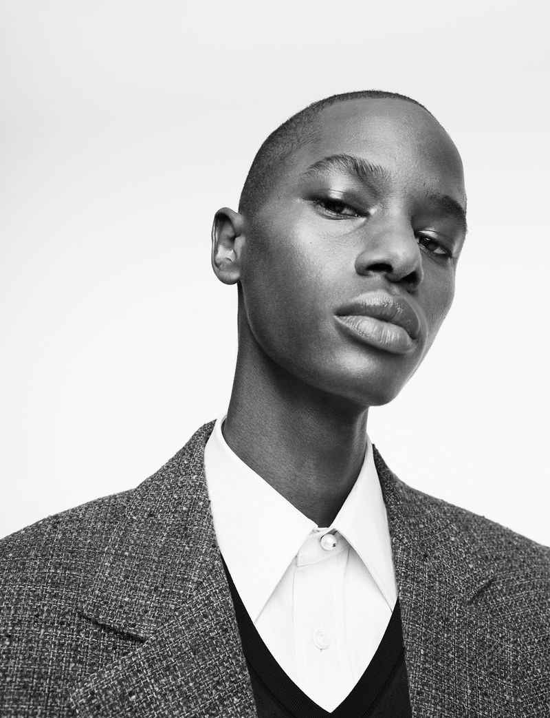 Close-up portrait of a young Black adult in a grey tweed blazer over a white shirt and V-neck sweater, against a neutral background.