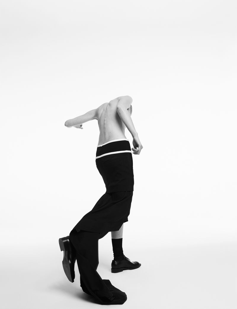 Monochrome studio shot of a human figure with partial clothing; black dress pants being removed, black shoes, and black socks.