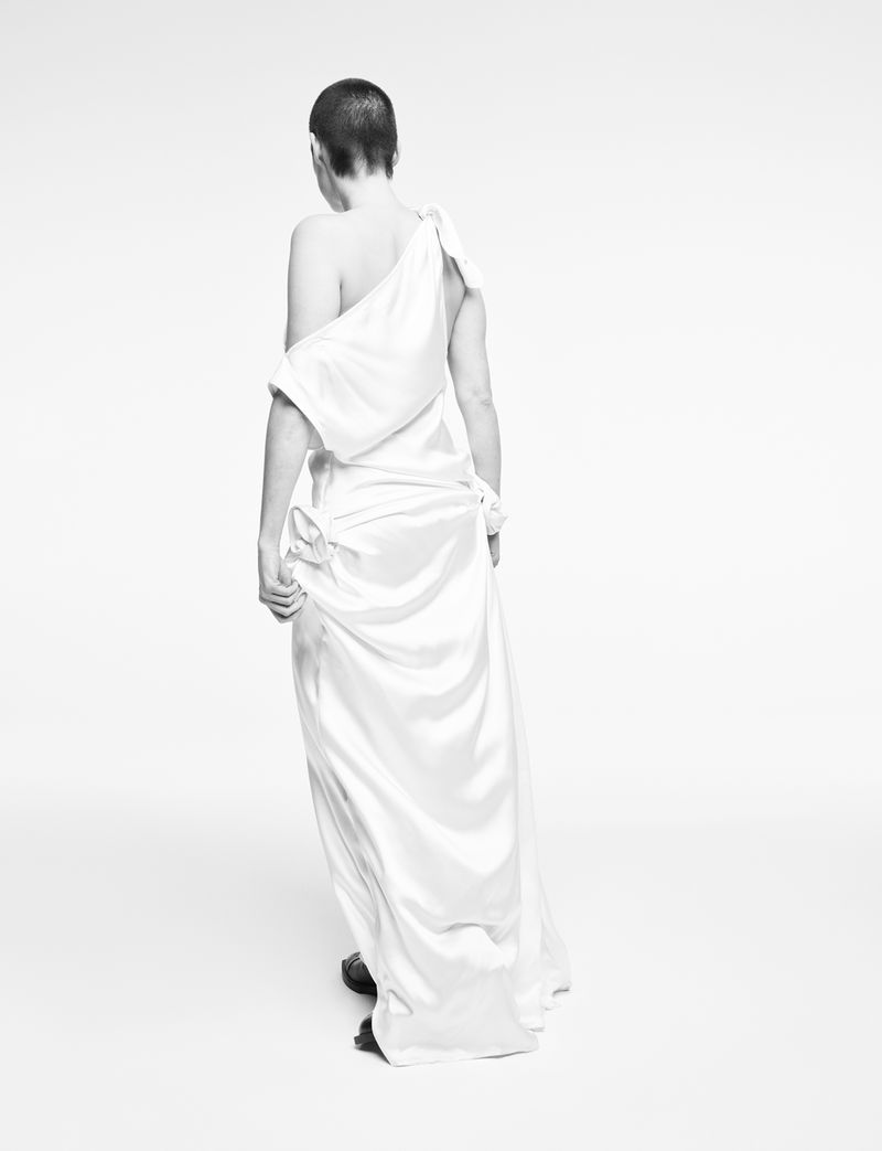 Back view of a woman with short hair wearing an elegant white dress in a bright studio setting, conveying simplicity and grace.