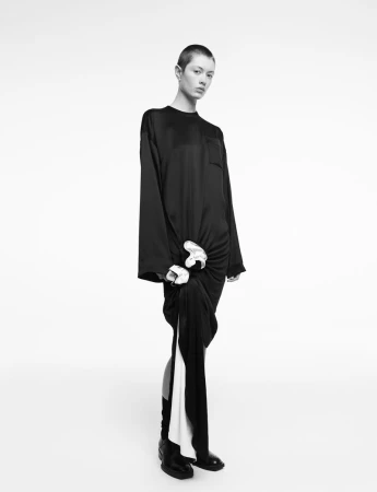 Model with a shaved head wearing a long black silk dress against a plain white seamless studio background.
