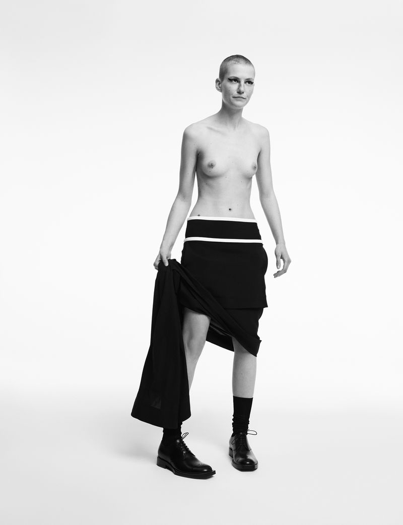 Monochrome studio shot of a shaved-head woman wearing a black skirt, boots, and holding a garment against a white backdrop.