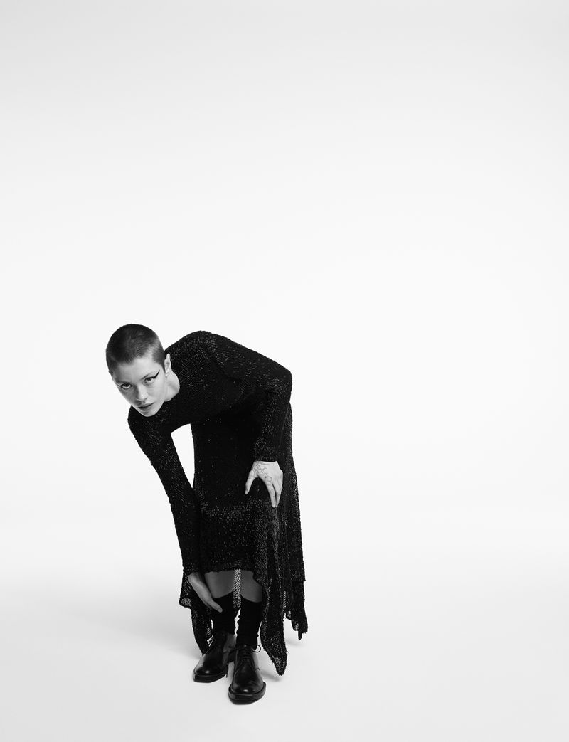 Woman in a sparkly long-sleeve dress and boots is bent forward slightly while posing in a photo studio against a white backdrop.