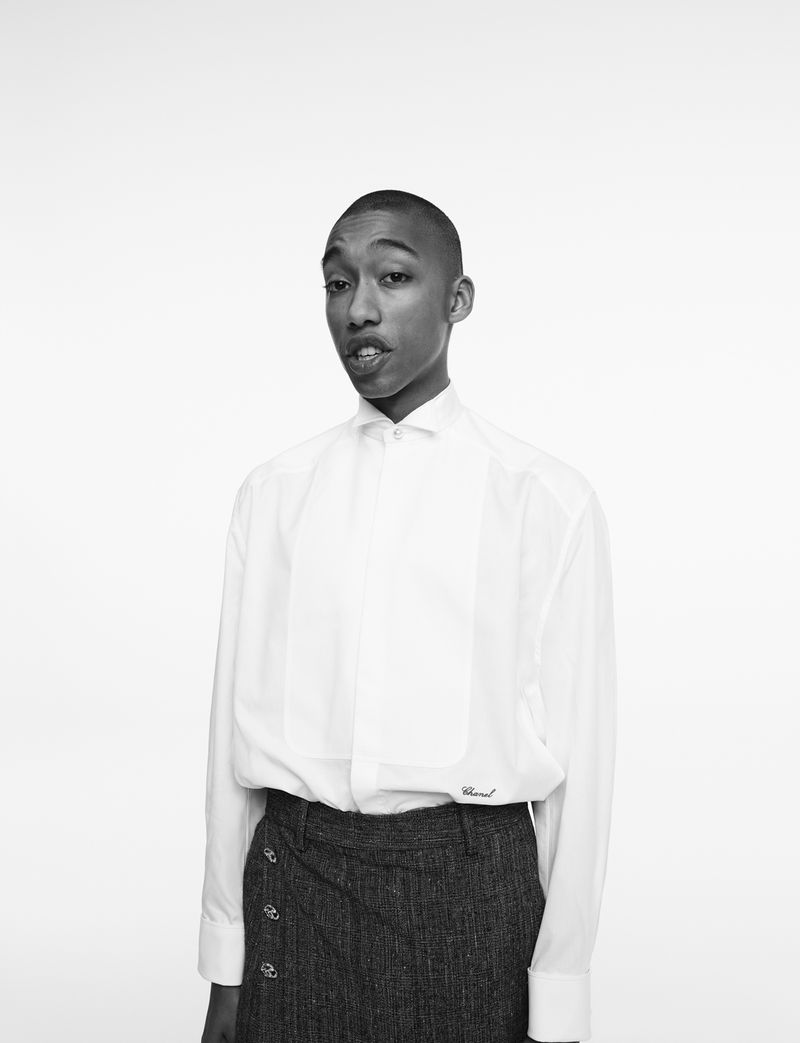 Portrait of a person with short hair wearing a white button-down shirt and dark grey tweed skirt against a white background.