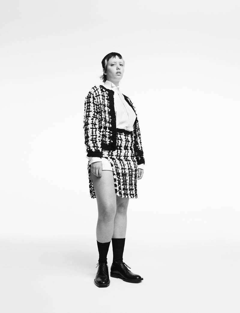 Full-length portrait of a woman in a tweed jacket and skirt with black socks and boots, posing against a white background in a studio.