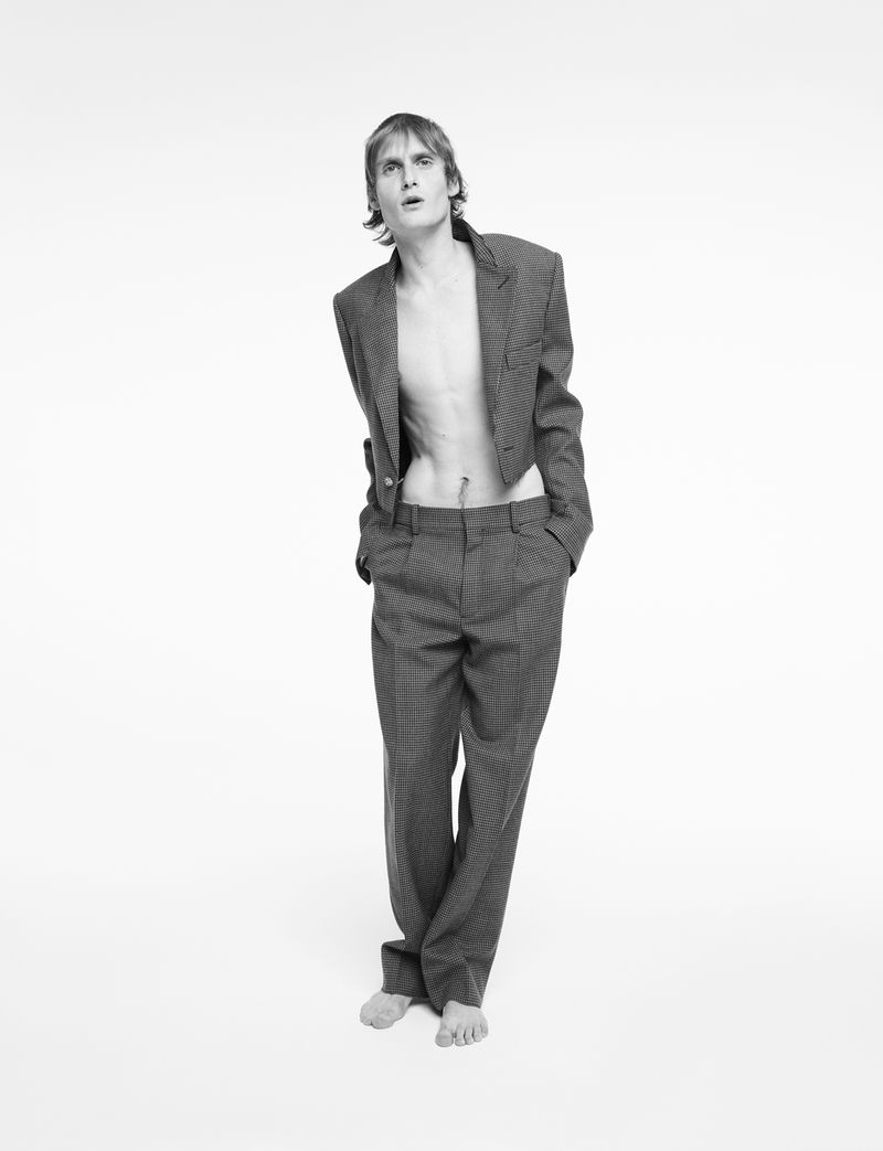 Monochrome studio portrait of a slender, light-skinned male fashion model in a cropped blazer and pants, hands in pockets, gazing upward.