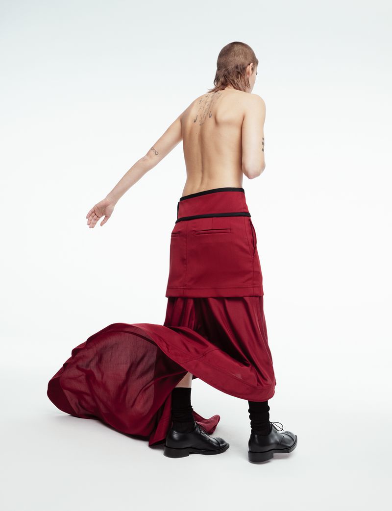 Person wearing a burgundy skirt and black socks and shoes stands in a white studio, back facing the camera.