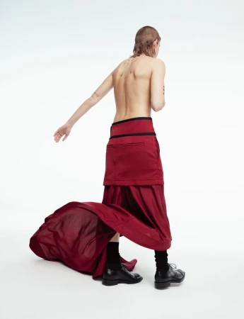 Person wearing a burgundy skirt and black socks and shoes stands in a white studio, back facing the camera.