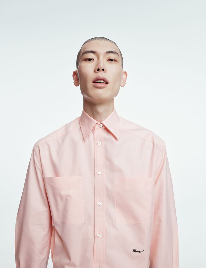 Portrait of a young adult male with a shaved head wearing a pink button-down shirt against a white background, fashion style.