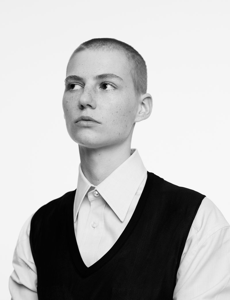Black and white portrait of a young adult female with a shaved head, wearing a button-down shirt and vest against a bright white background.