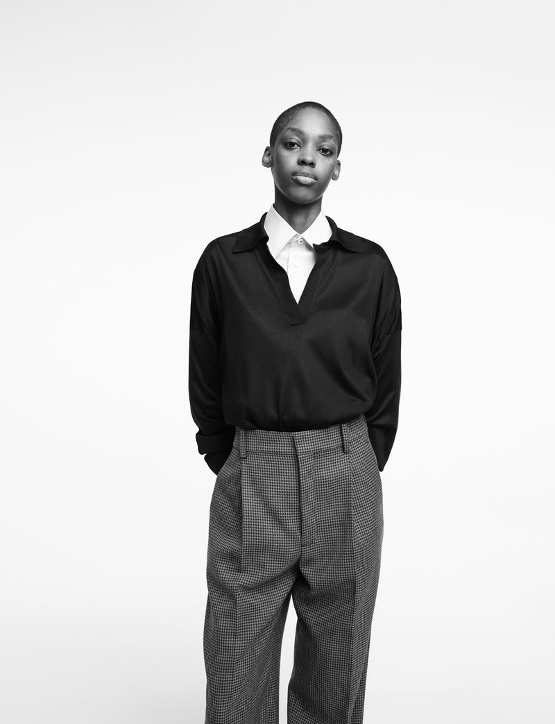 Black-and-white portrait of an androgynous person with short hair wearing a dark top and patterned pants against a bright backdrop.