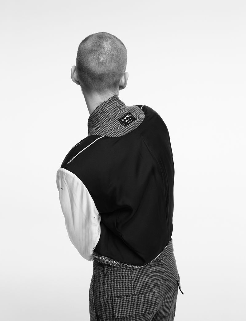 Back view of a slim adult man in a stylish black and grey jacket and grey checkered pants against a white backdrop.