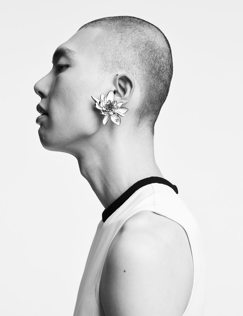 Monochrome studio portrait of a short-haired Asian man in a sleeveless shirt wearing a flower earring against a white backdrop.