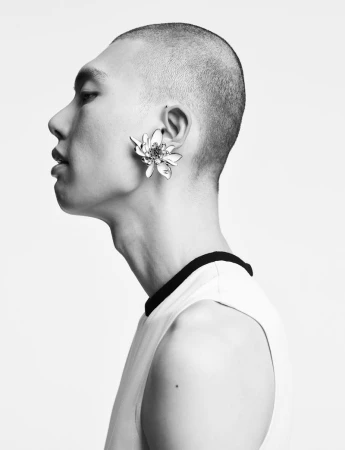 Monochrome studio portrait of a short-haired Asian man in a sleeveless shirt wearing a flower earring against a white backdrop.