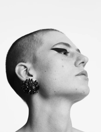 A close-up profile portrait of a person with a shaved head and bold black winged eyeliner wearing a large, textured black floral earring.