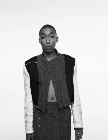 A person with a shaved head wearing a dark textured cropped jacket and high-waisted trousers against a plain white studio background.