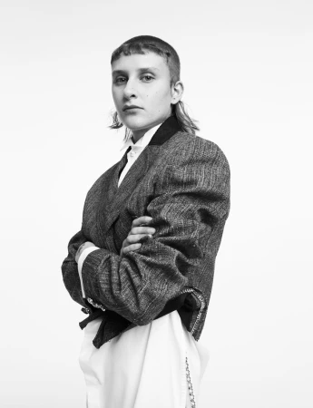 Black and white portrait of a person with short hair wearing a tweed blazer, posing with arms crossed against a white backdrop.