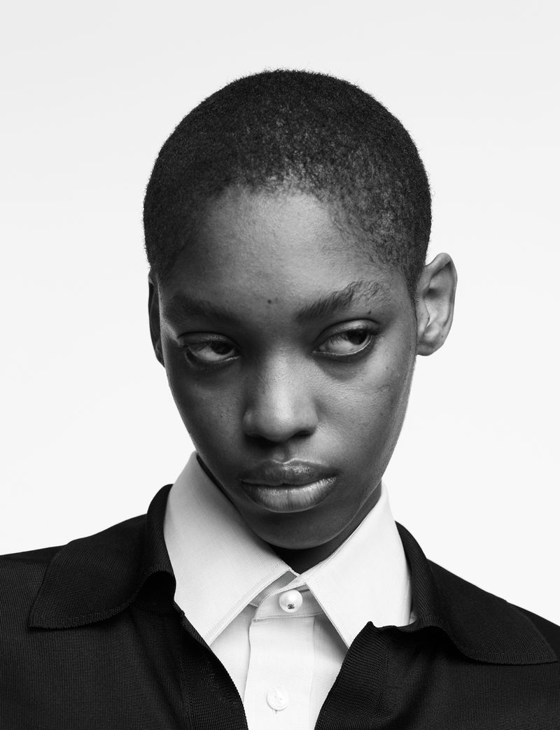 Monochromatic portrait of a young Black woman in a collared shirt against a white backdrop, looking off to the side in a minimalist composition.