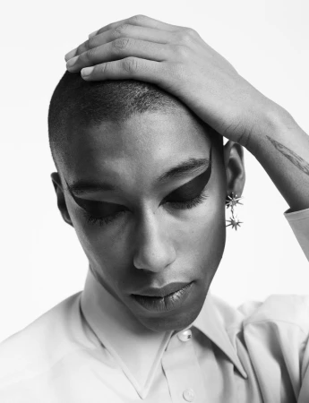 Close-up of a person with shaved hair, graphic black winged eyeliner, and star-shaped earrings, holding their head with one hand.
