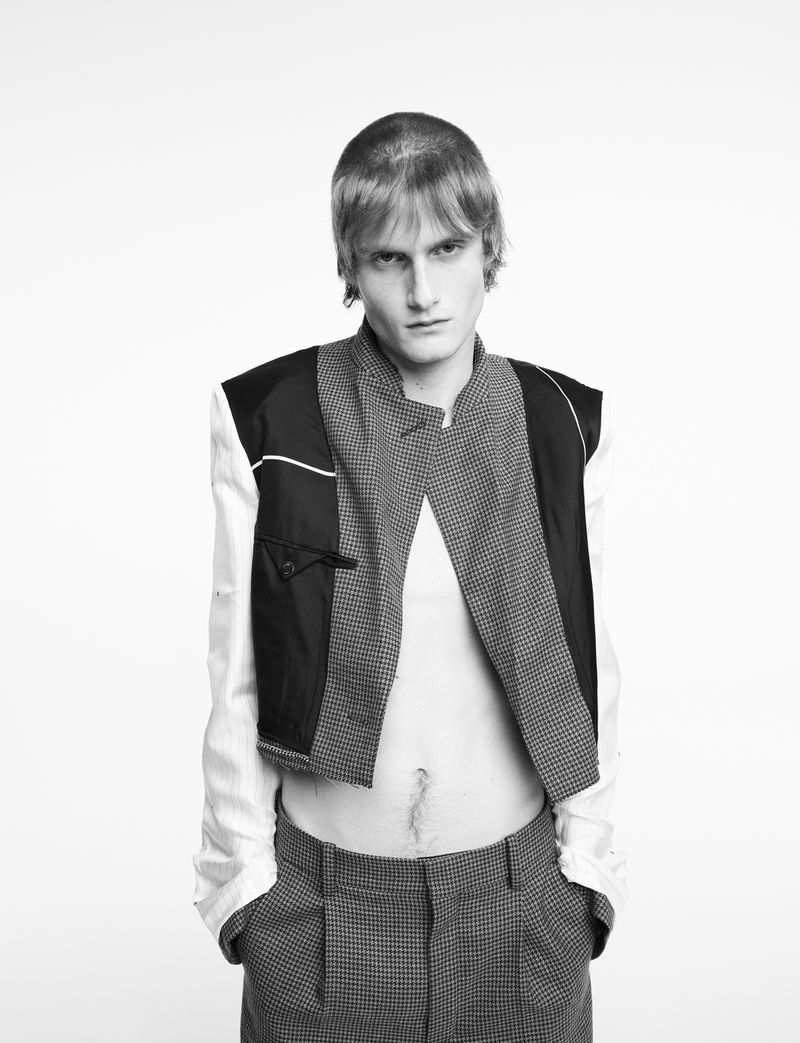 Black-and-white studio portrait of a fair-skinned, slender male model with a serious expression wearing avant-garde fashion with houndstooth pants.