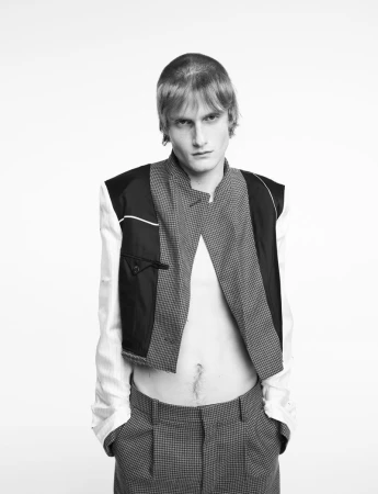 Black-and-white studio portrait of a fair-skinned, slender male model with a serious expression wearing avant-garde fashion with houndstooth pants.