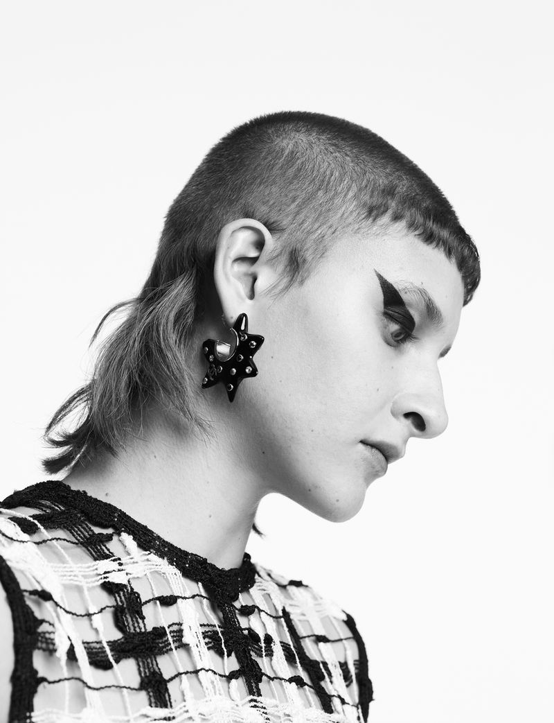 Close-up black and white portrait of a model wearing a statement star earring and a sheer top in a photo studio with a white backdrop.