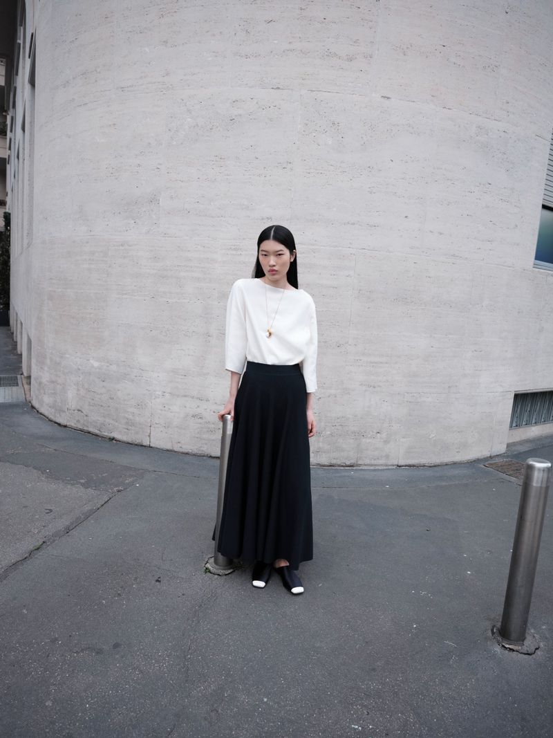 Elegant Asian woman in a white blouse and black skirt poses against a concrete wall on a city street. Minimalist fashion portrait.