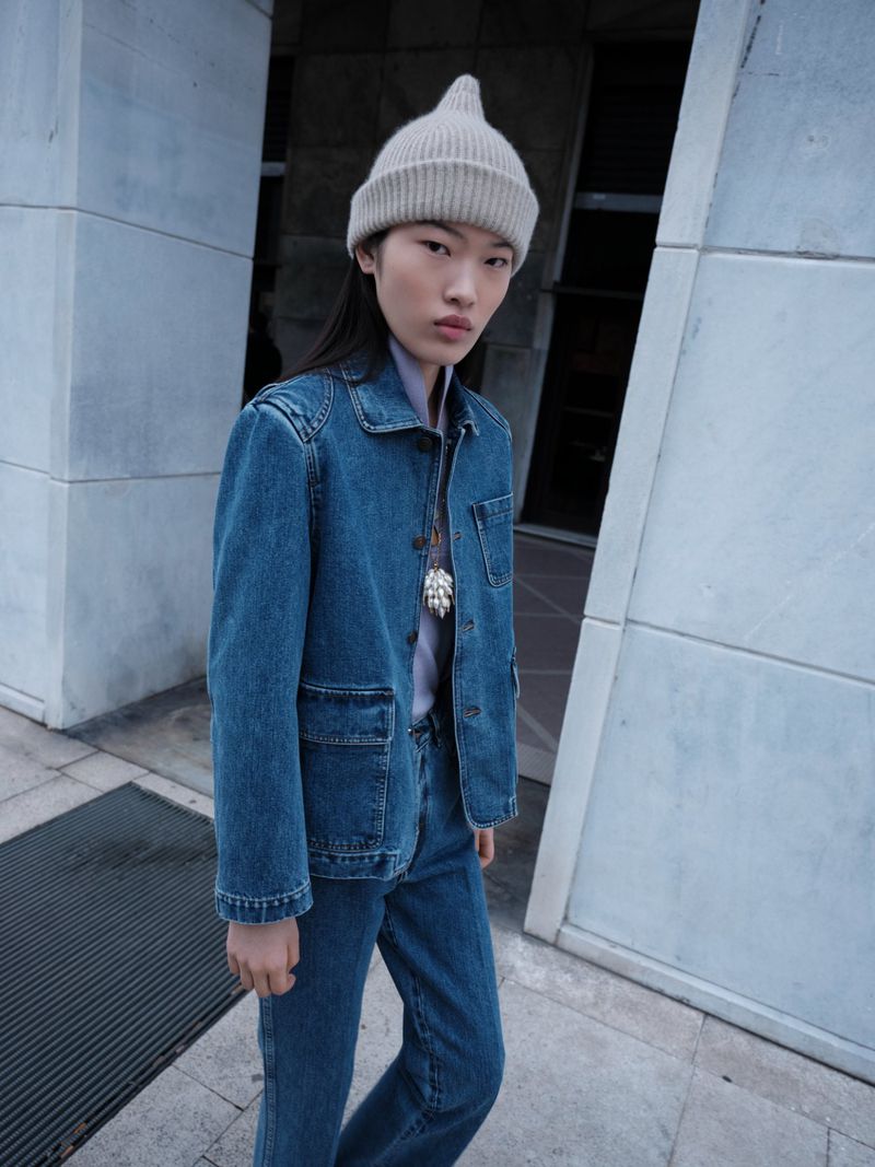 Fashion portrait of a young adult Asian woman in a blue denim jacket and jeans, posing against an exterior urban building.