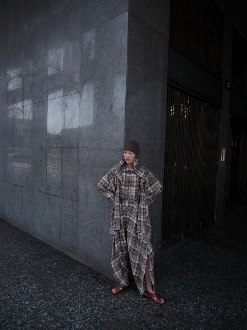 Woman in a plaid outfit with a hat and brown leather sandals stands against a concrete wall in front of a building entrance.