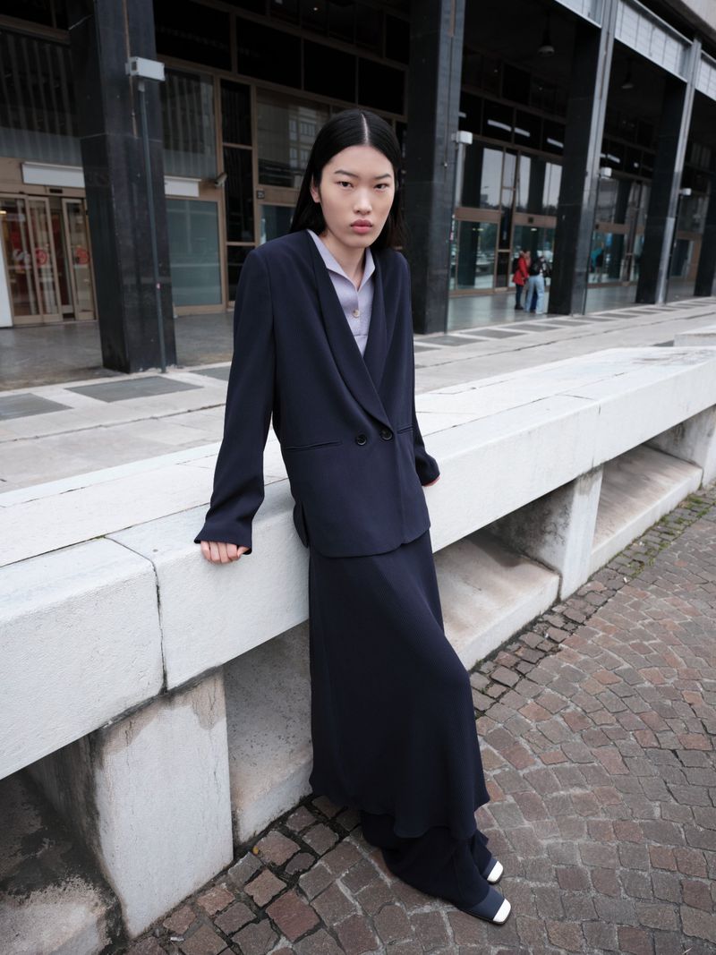 Asian woman in a dark suit and skirt leans against a concrete wall outdoors. Editorial fashion shot with natural light.