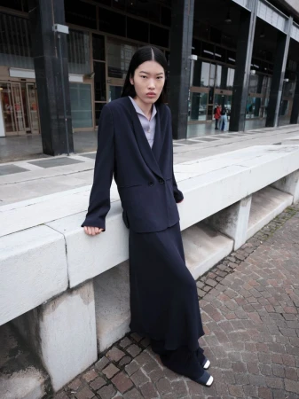 Asian woman in a dark suit and skirt leans against a concrete wall outdoors. Editorial fashion shot with natural light.