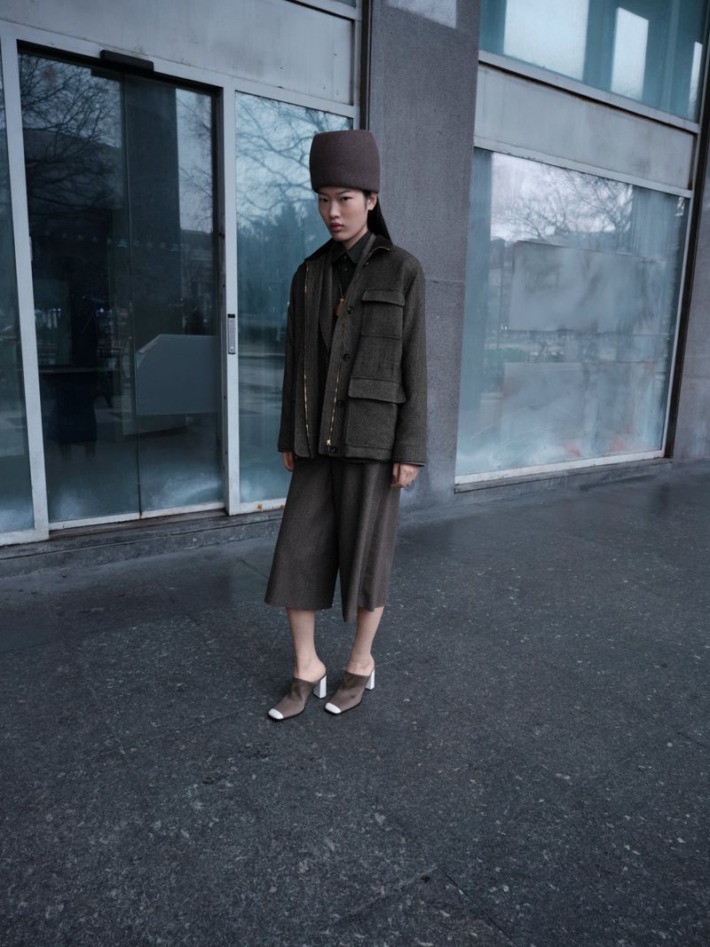 Asian woman poses outdoors wearing a brown jacket, wide-leg culottes, clogs, and a pillbox hat in an urban setting near a glass building.