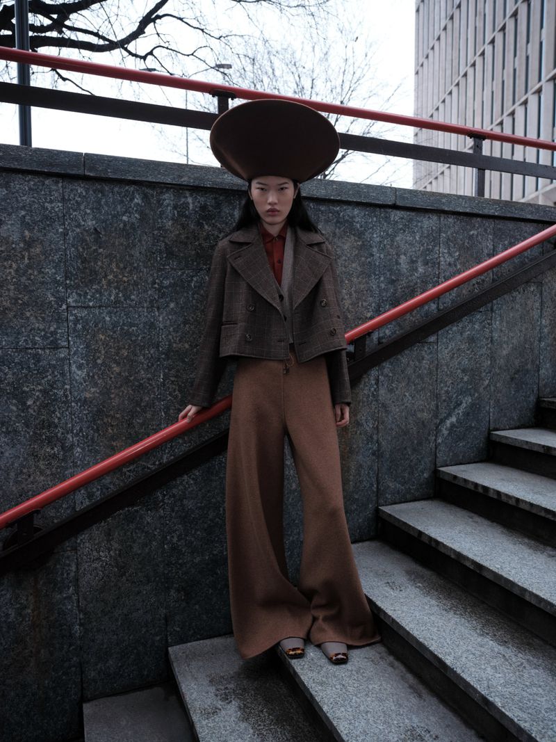 A woman in brown wide-leg pants and a blazer poses on outdoor stairs, wearing a large hat; a modern building is visible in the background.