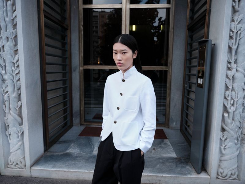 An Asian woman stands outdoors in a white buttoned jacket and black pants, posing in front of a detailed stone building entrance.