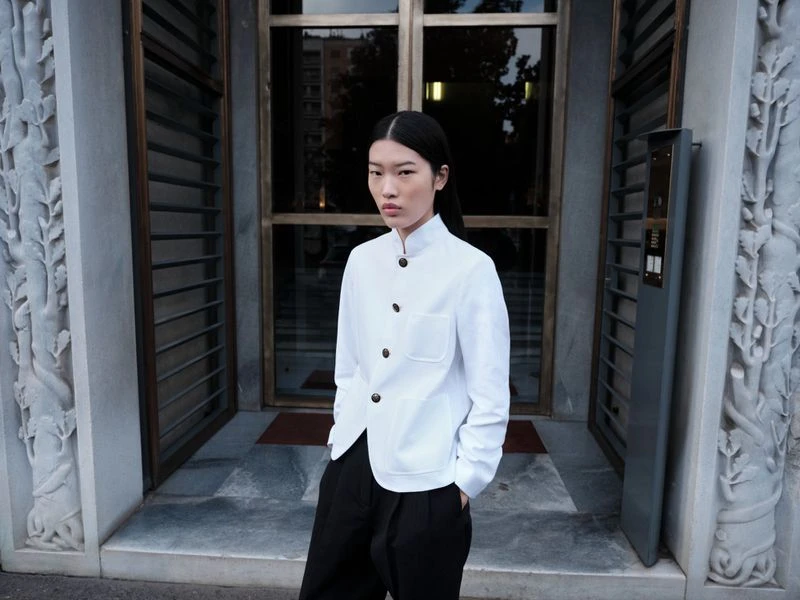 An Asian woman stands outdoors in a white buttoned jacket and black pants, posing in front of a detailed stone building entrance.