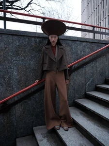 Model wearing a wide-brimmed brown hat, tweed jacket, and wide trousers standing on stone outdoor stairs with a red handrail.