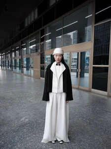 Model standing outdoors on stone pavement in front of a modern glass and metal building wearing a white hat, black and white jacket, and wide-leg trousers