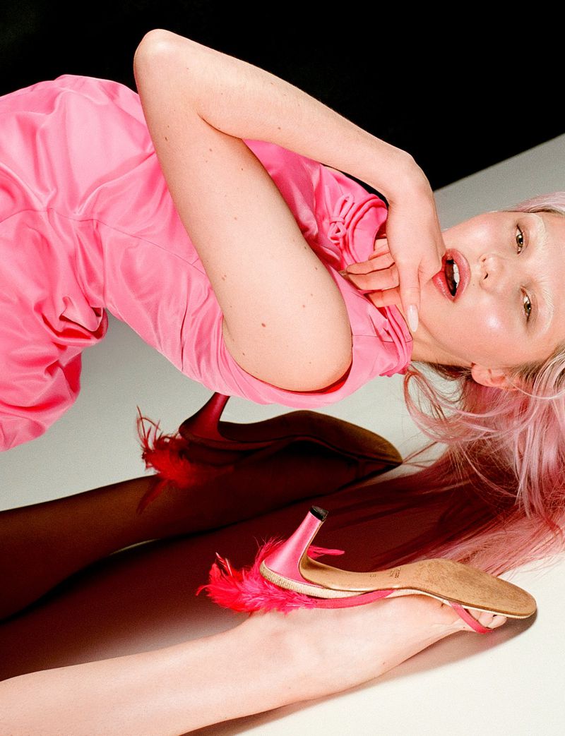 Young woman lying down in a pink dress with pink hair and pink heels with feather detail, against a black and white background.