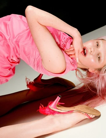 Young woman lying down in a pink dress with pink hair and pink heels with feather detail, against a black and white background.