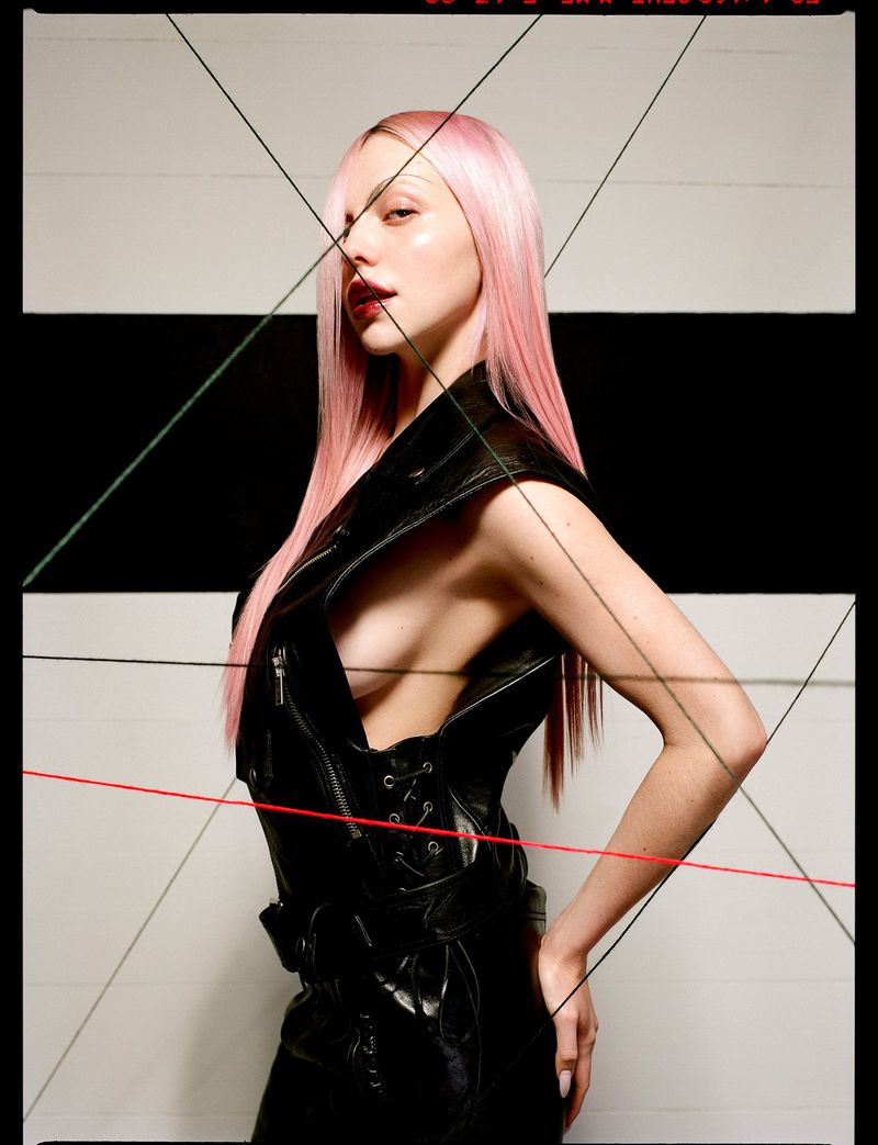 A woman with pink hair poses in a black vest with geometric string art, against a backdrop of white and black stripes.